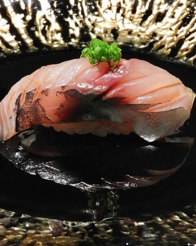 A single piece of seared nigiri sushi, glistening, on a black plate with a gold rim.