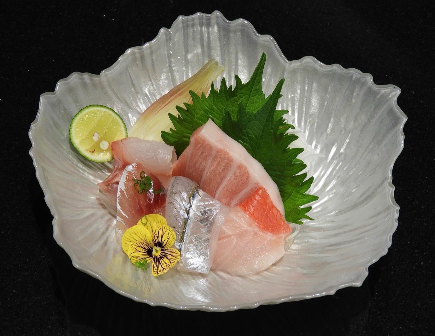 An assortment of sashimi on a shell-shaped glass plate, with shiso, citrus and an edible flower.