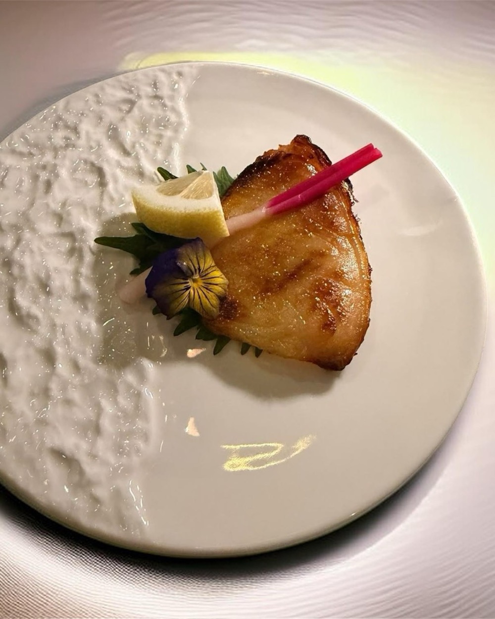 A seared piece of toothfish, plated with a citrus wedge and edible flowers.